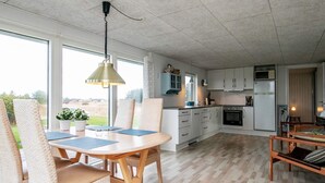 Une cuisine moderne avec des meubles blancs, un coin repas avec une table en bois et des chaises en osier, et une vue sur l’extérieur grâce à de grandes fenêtres.
