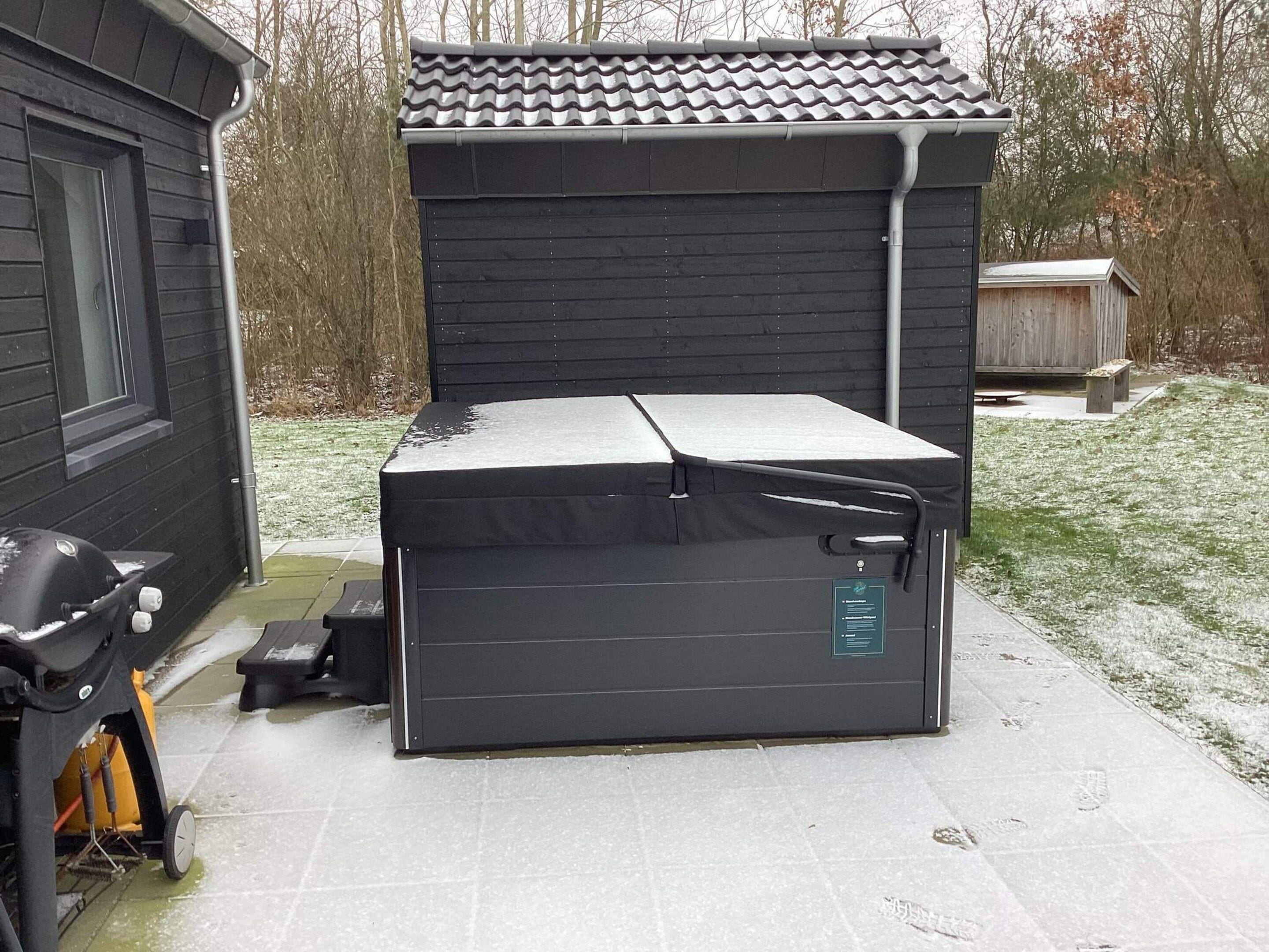 A compact, dark-colored outdoor storage unit with a flat top, possibly a shed or a small garage, situated on a paved area with a grassy yard in the background.