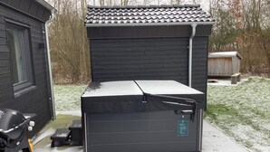 Un depósito exterior compacto y de color oscuro con tapa plana, posiblemente un cobertizo o un pequeño garaje, ubicado en una zona pavimentada con un césped al fondo.