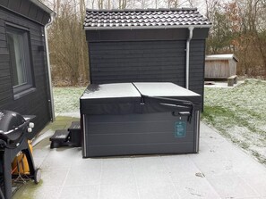 En lille, mørk udendørs opbevaringsenhed med fladt tag, måske et skur eller et lille garage, placeret på en belagt flade med en græsplæne i baggrunden.