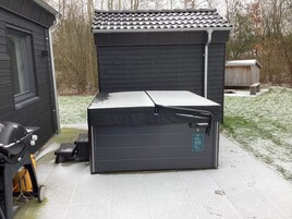 Un depósito exterior compacto y de color oscuro con tapa plana, posiblemente un cobertizo o un pequeño garaje, ubicado en una zona pavimentada con un césped al fondo.