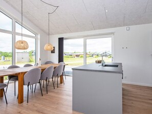 A modern kitchen with a large island, a dining area with a wooden table and grey chairs, and large windows offering a view of the outdoors.
