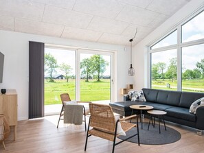A spacious living room with a large sofa, two wicker chairs, a small round table, and a view of a green landscape through large windows. - 5 Star Holiday Home in Ansager (Ansager)