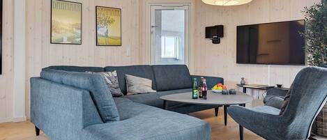 A modern living room with a sectional sofa, a round coffee table, and a single armchair. There is a flat-screen TV mounted on the wall and a large window with a view of the outdoors.