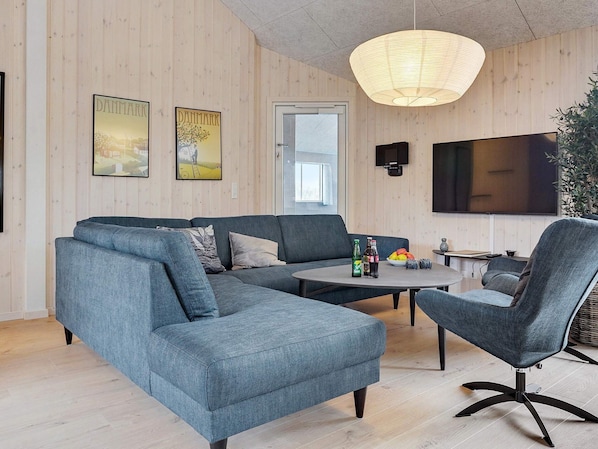 A modern living room with a sectional sofa, a round coffee table, and a single armchair. There is a flat-screen TV mounted on the wall and a large window with a view of the outdoors.