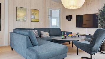Een moderne woonkamer met een hoekbank, een ronde salontafel en een fauteuil. Een televisie is aan de muur bevestigd, er is een deur en twee ingelijste foto’s hangen aan de muur.