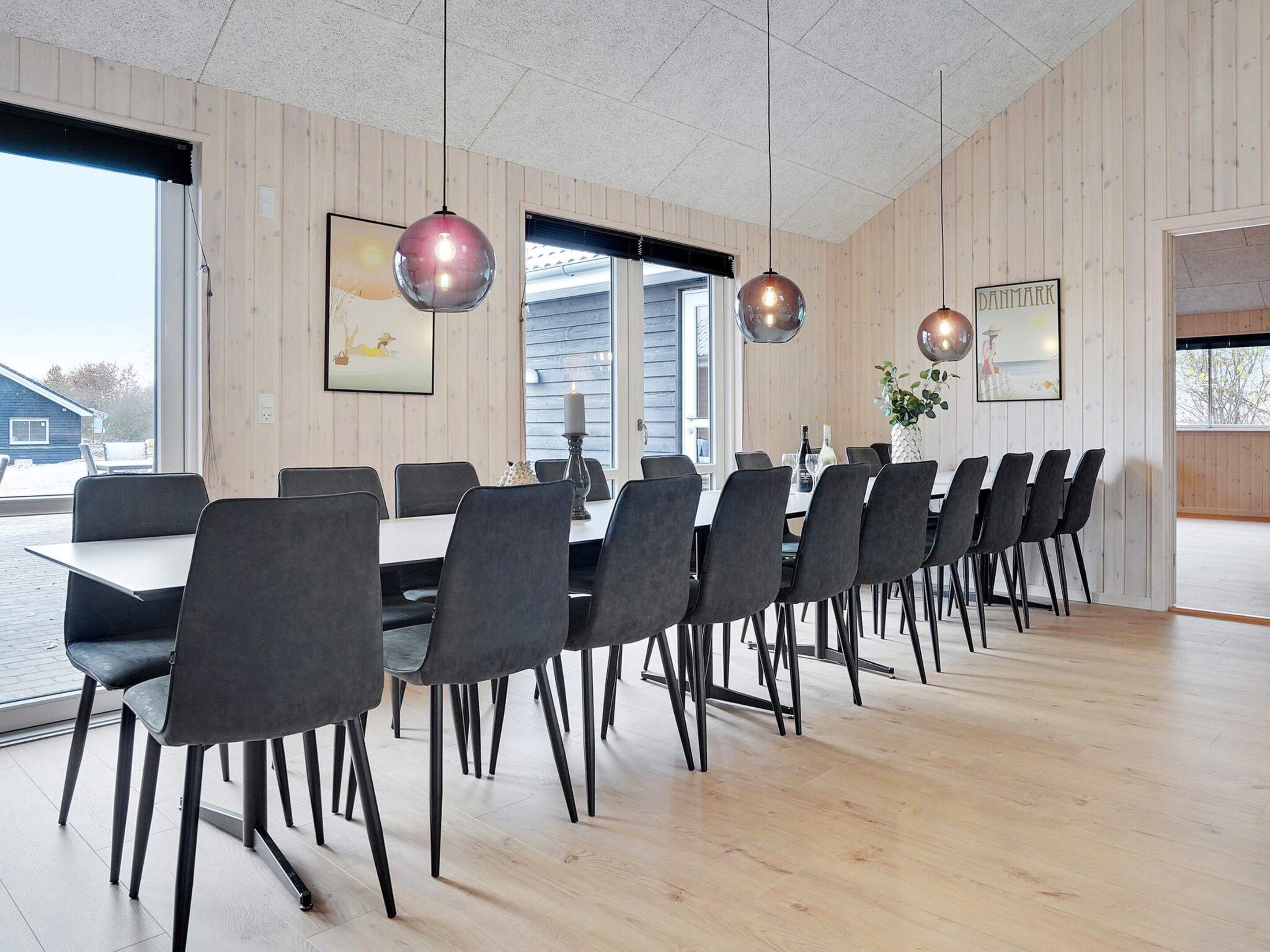 Ein moderner Essbereich mit einem langen Tisch, mehreren Stühlen, Pendelleuchten und Blick ins Freie.