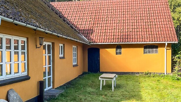 A yellow building with a red-tiled roof, a small white table, and a grassy yard with rocks. - 5 Person Holiday Home in Skanderborg (Skanderborg)