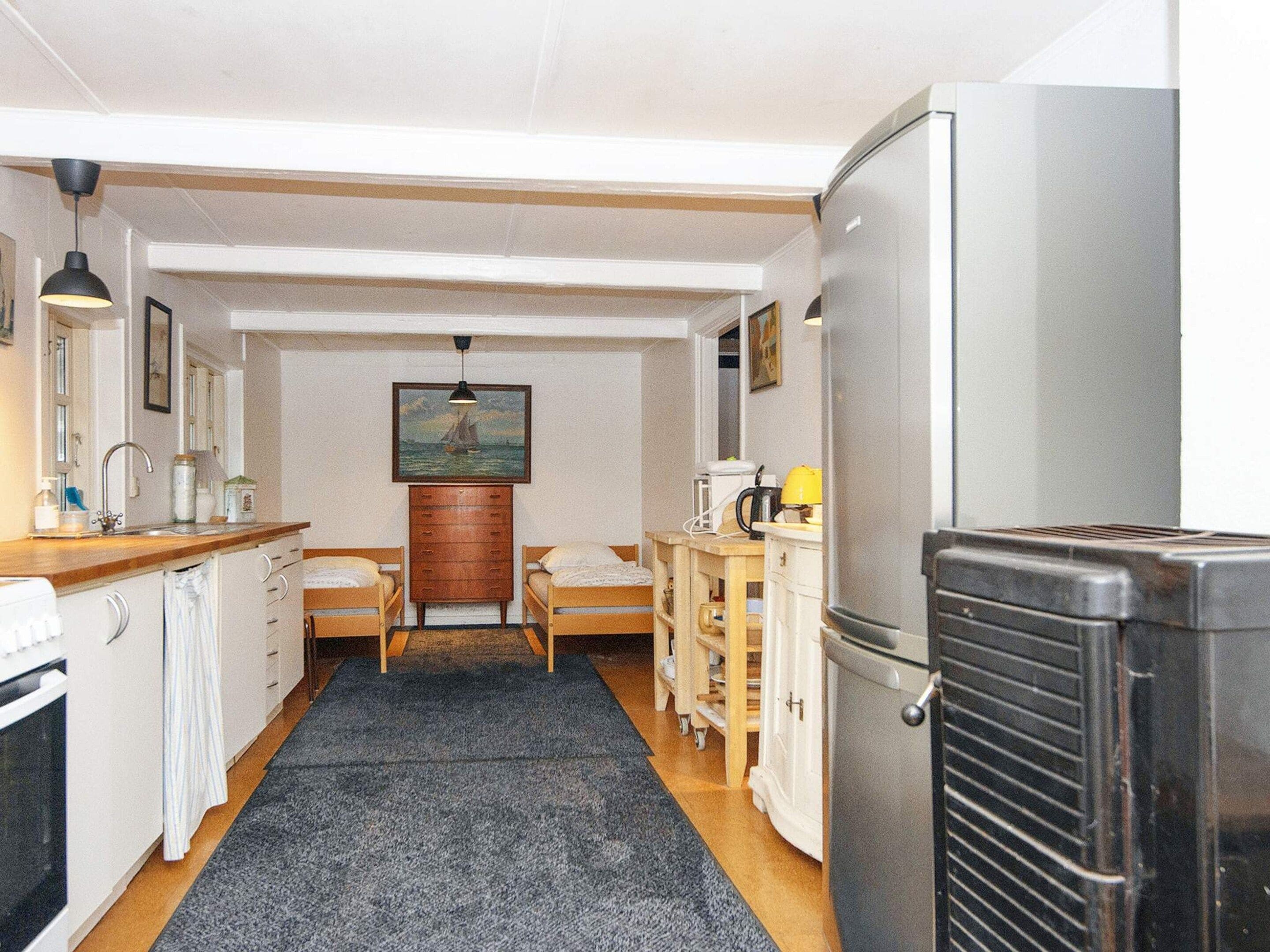 A compact kitchen with a refrigerator, sink, and wooden cabinets. There are two beds with wooden frames and a painting on the wall.