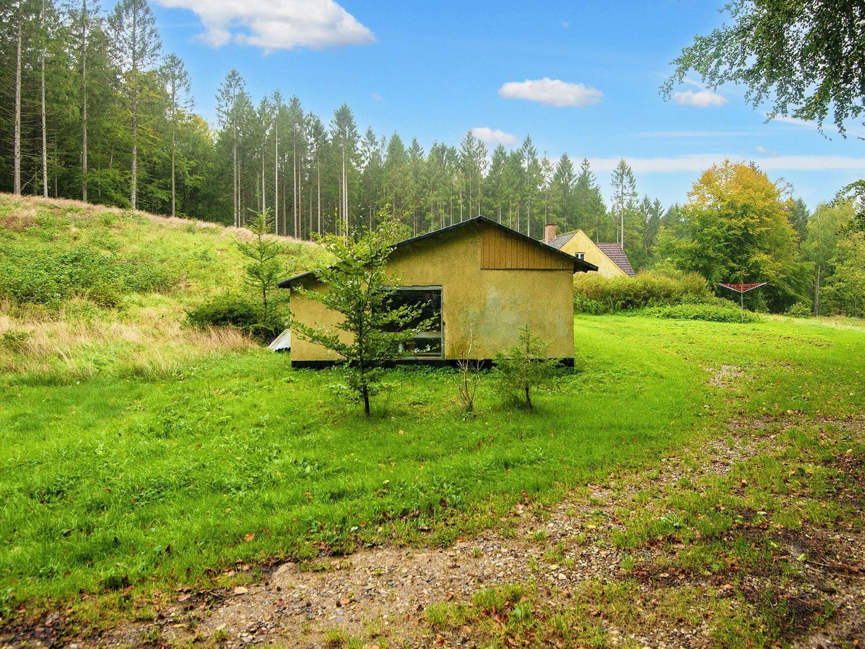 A small wooden cabin with a sloped roof, surrounded by a grassy field and dense forest.