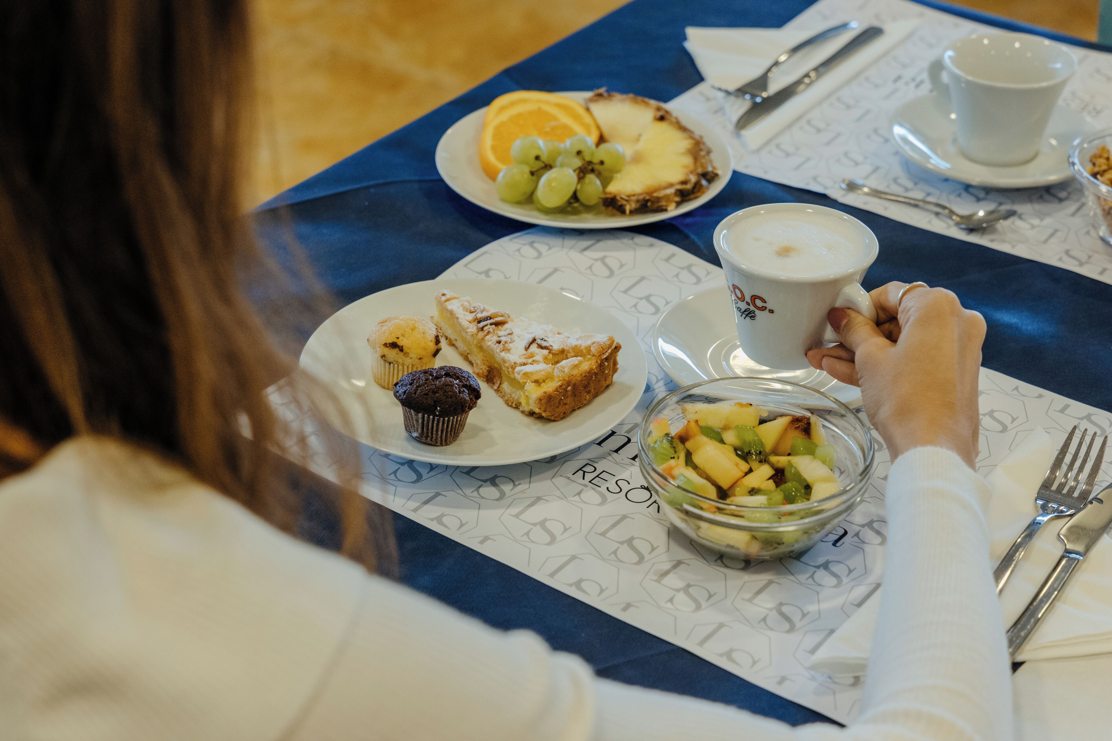 Ein Esstisch mit blauer Tischdecke, Tellern mit Speisen, einer Schüssel mit Obstsalat und einer Tasse Kaffee.