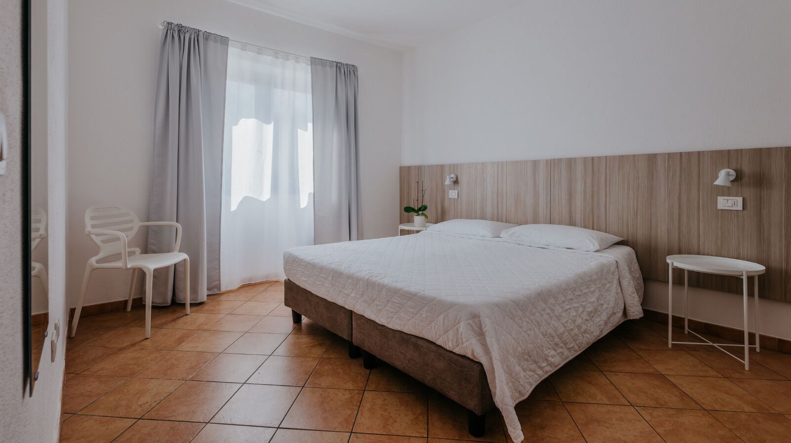 Een slaapkamer met een bed, nachtkastje, stoel en raam met gordijnen.