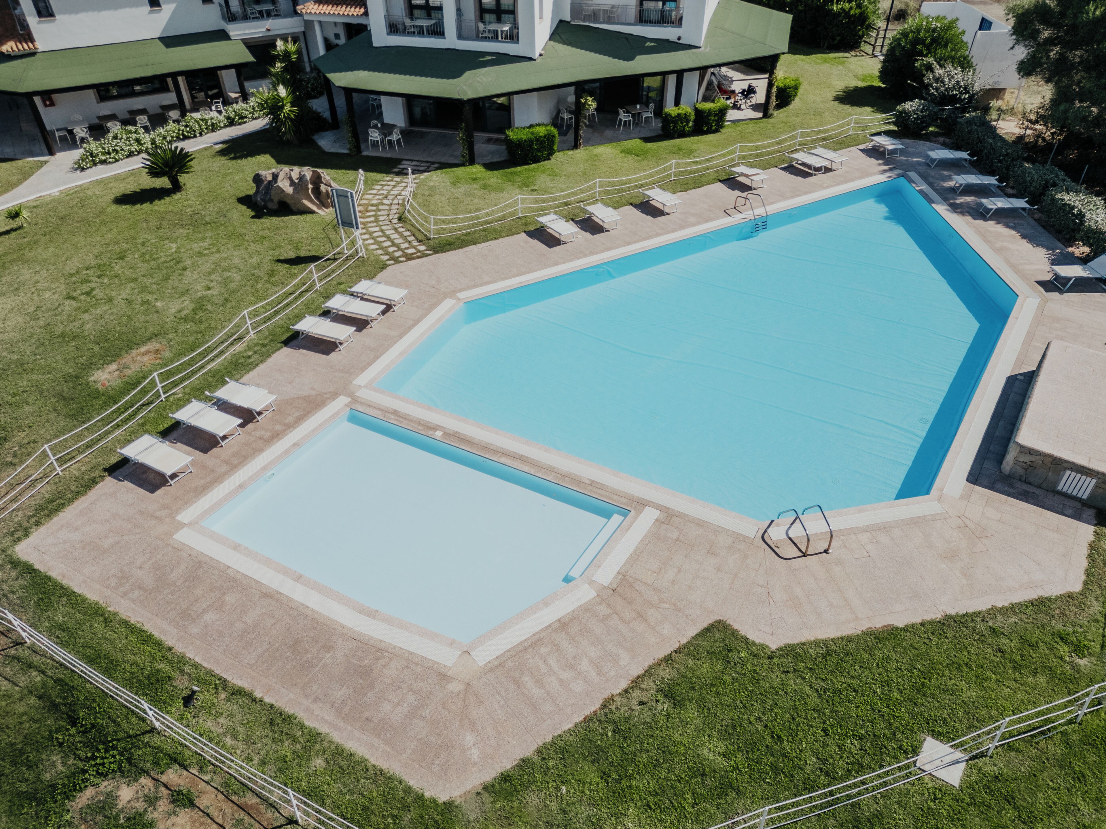Une grande piscine rectangulaire, avec une piscine plus petite attenante, entourée d’une terrasse en béton et d’une zone herbeuse agrémentée de chaises longues.