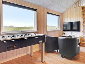 Una habitación con mesa de fútbol, dos ventanas con vista a un campo, televisor de pantalla plana y estantería de madera.