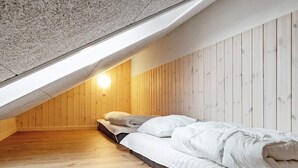 Quarto de sótão com teto inclinado, paredes de madeira, cama com roupa de cama branca e luminária.
