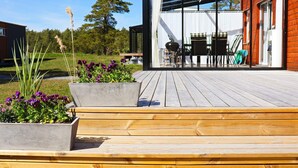 A wooden deck with concrete planters of purple flowers, a glass-enclosed patio area, and a view of a barbecue grill and outdoor furniture. - 6 Person Holiday Home in Saffle (Saffle)