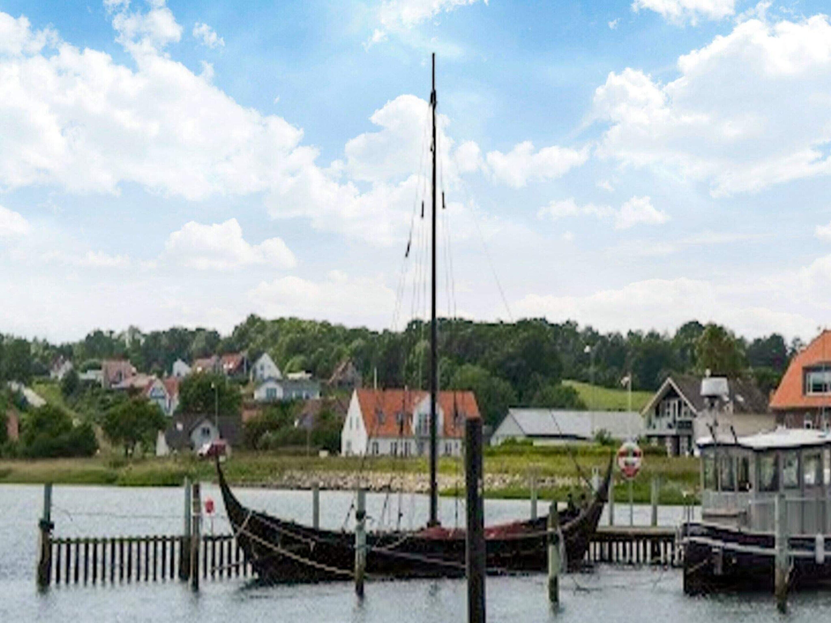 Een traditionele lange zeilboot afgemeerd aan een steiger, met op de achtergrond huizen en groen.