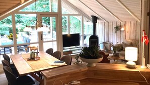 Une pièce spacieuse avec un plafond en bois, de grandes fenêtres, une cheminée, un téléviseur et un coin repas comprenant une table et des chaises.