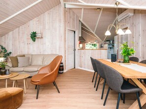 Une pièce chaleureuse et bien éclairée, avec un plafond en bois, un canapé, une table à manger avec des chaises, et un coin cuisine à l’arrière-plan.