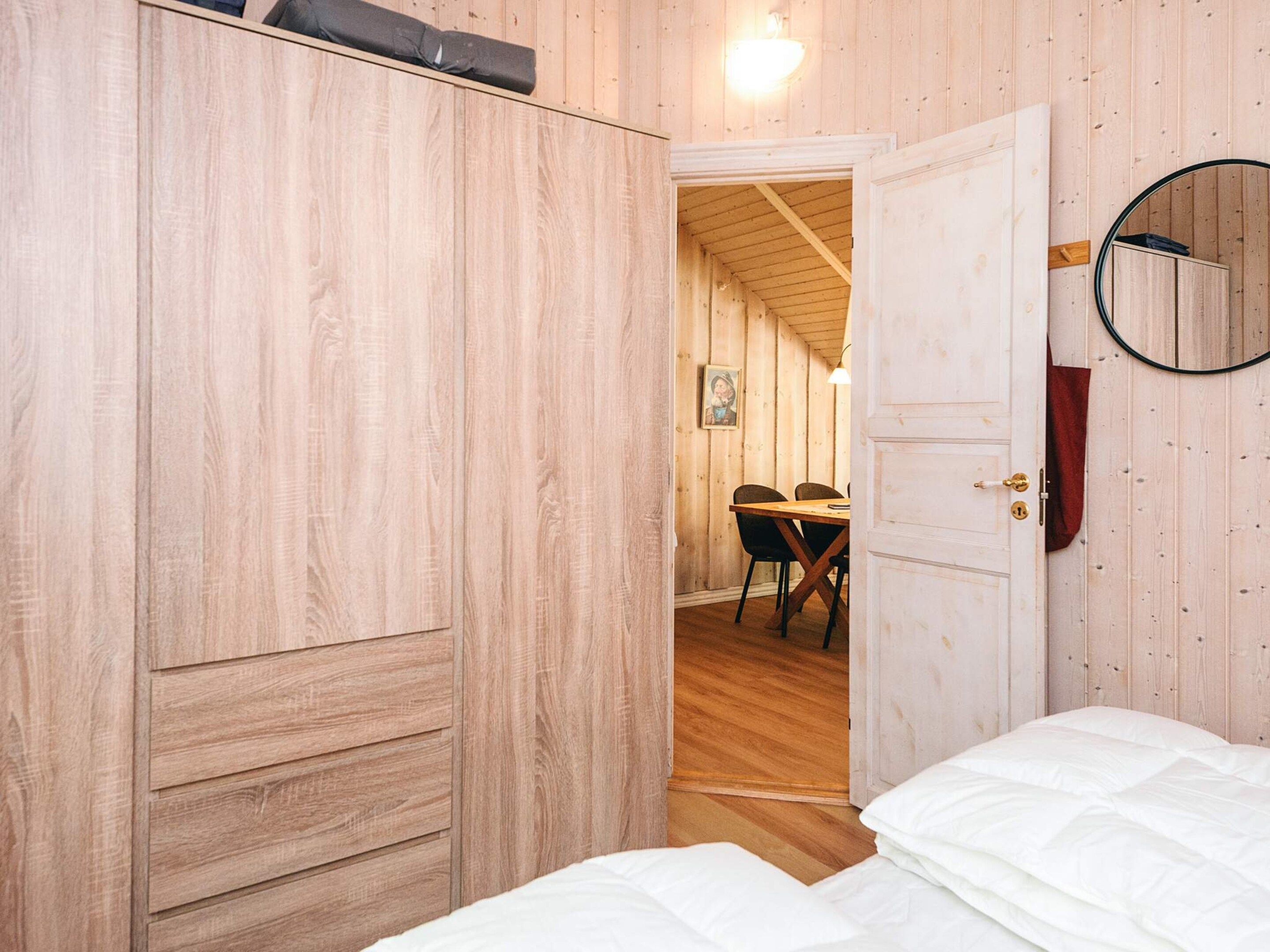 A wooden room with a bed, a door leading to a dining area, and a round mirror on the wall.