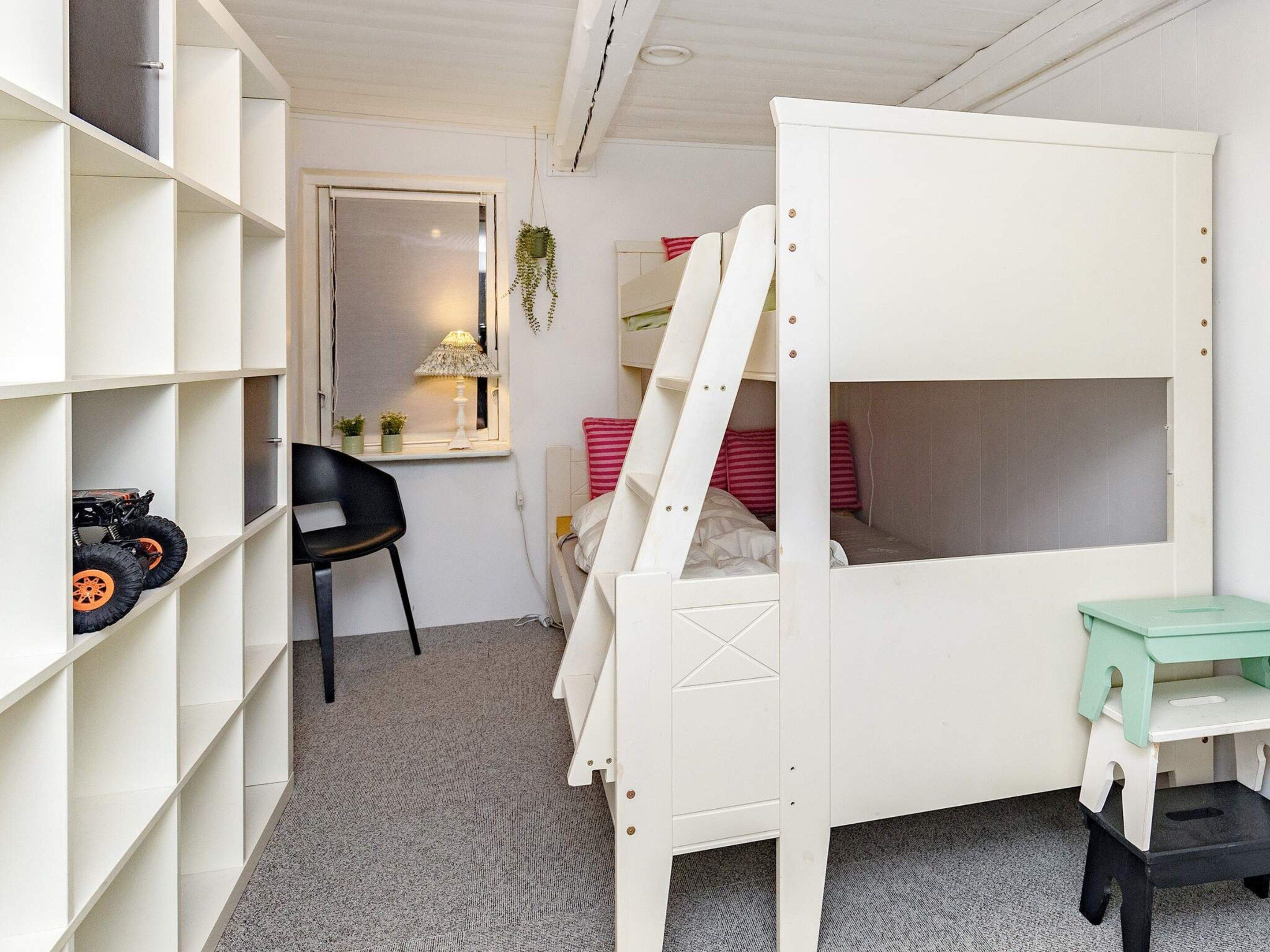A child's bedroom with a bunk bed, a desk, shelves, a chair, and a window with a lamp.