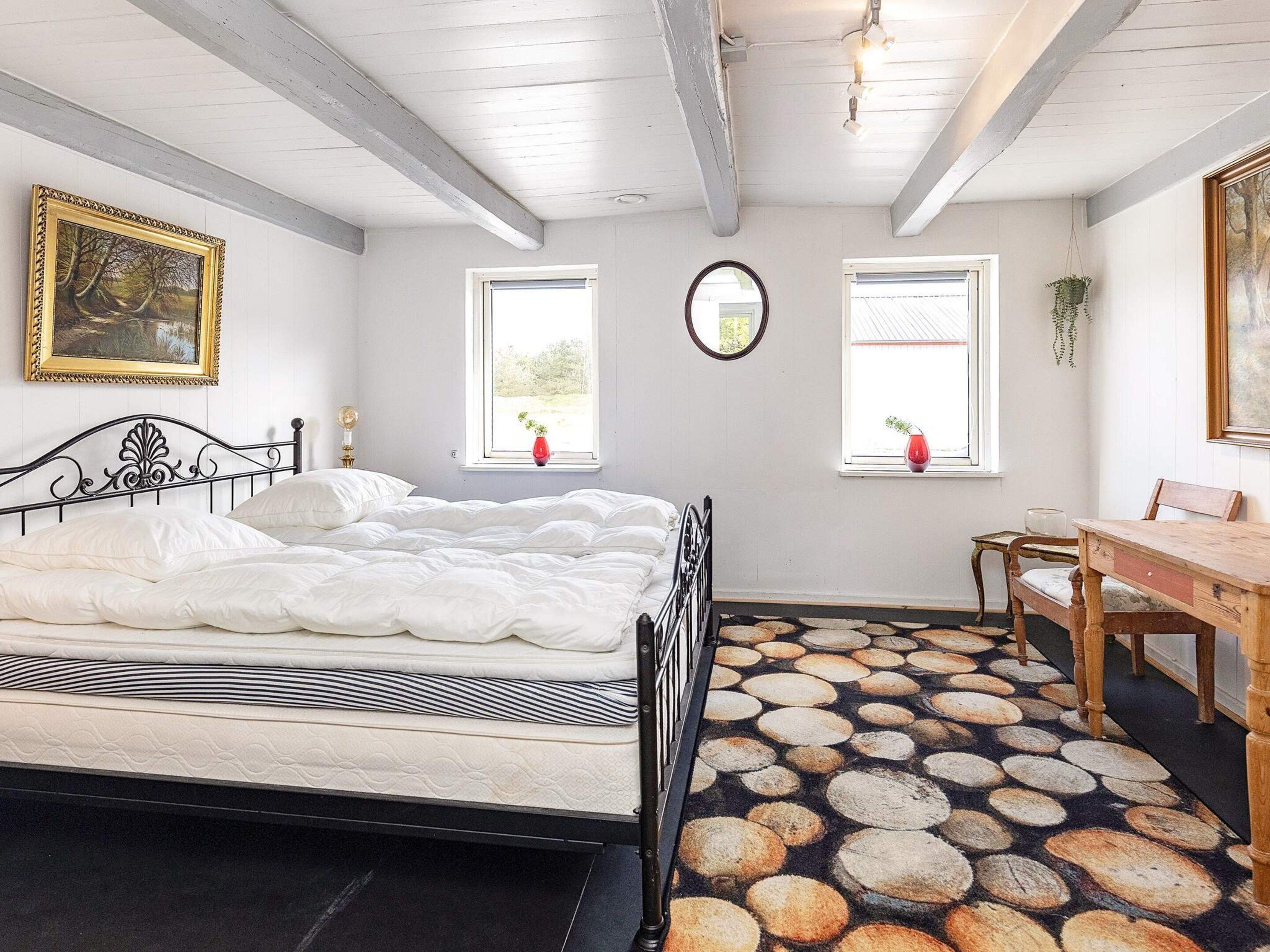 Une chambre à coucher avec un lit, deux fenêtres, un miroir, une table en bois et une chaise.
