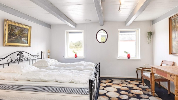 Un dormitorio con cama, dos ventanas, un espejo, una mesa de madera y una silla.