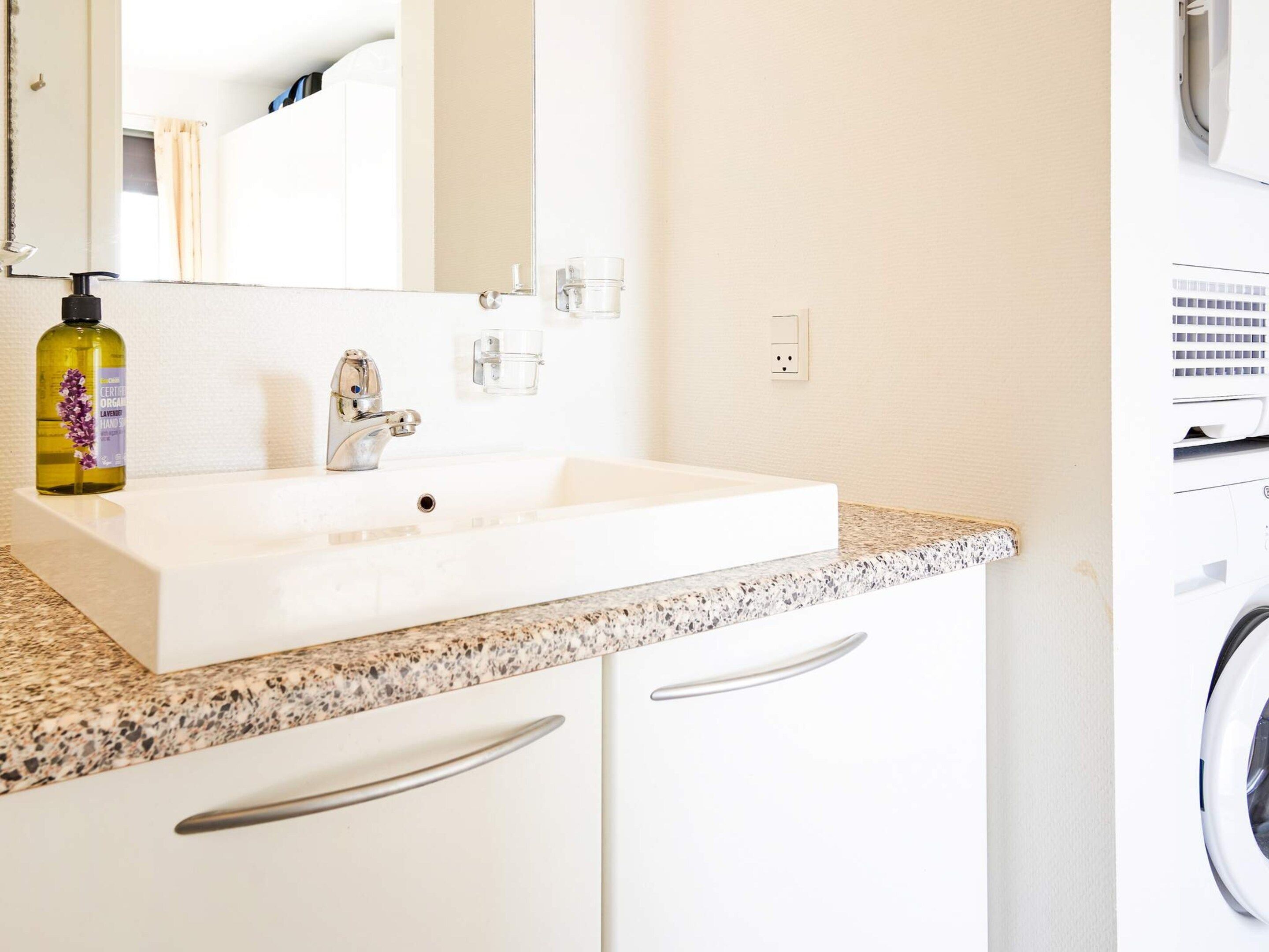 Une salle de bain avec un lavabo blanc, un miroir, un flacon de savon liquide et un lave-linge.