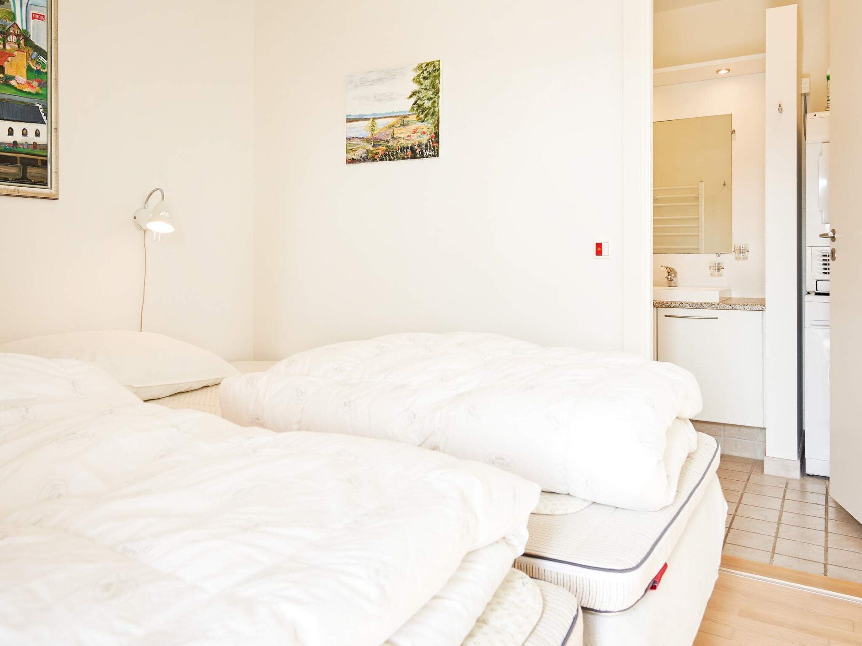 Une chambre d’hôtel avec deux lits, une table de chevet, une lampe fixée au mur, un petit tableau et une vue sur la cuisine adjacente.