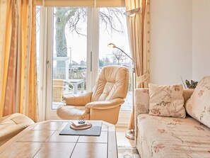 A living room with a leather chair, a sofa with floral cushions, a coffee table with a book and a decorative plate, and a view through a glass door.