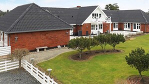 A two-story brick building with a balcony, a covered patio, and a fenced yard with trees. - 6 Person Holiday Home in Blavand (Blavand)