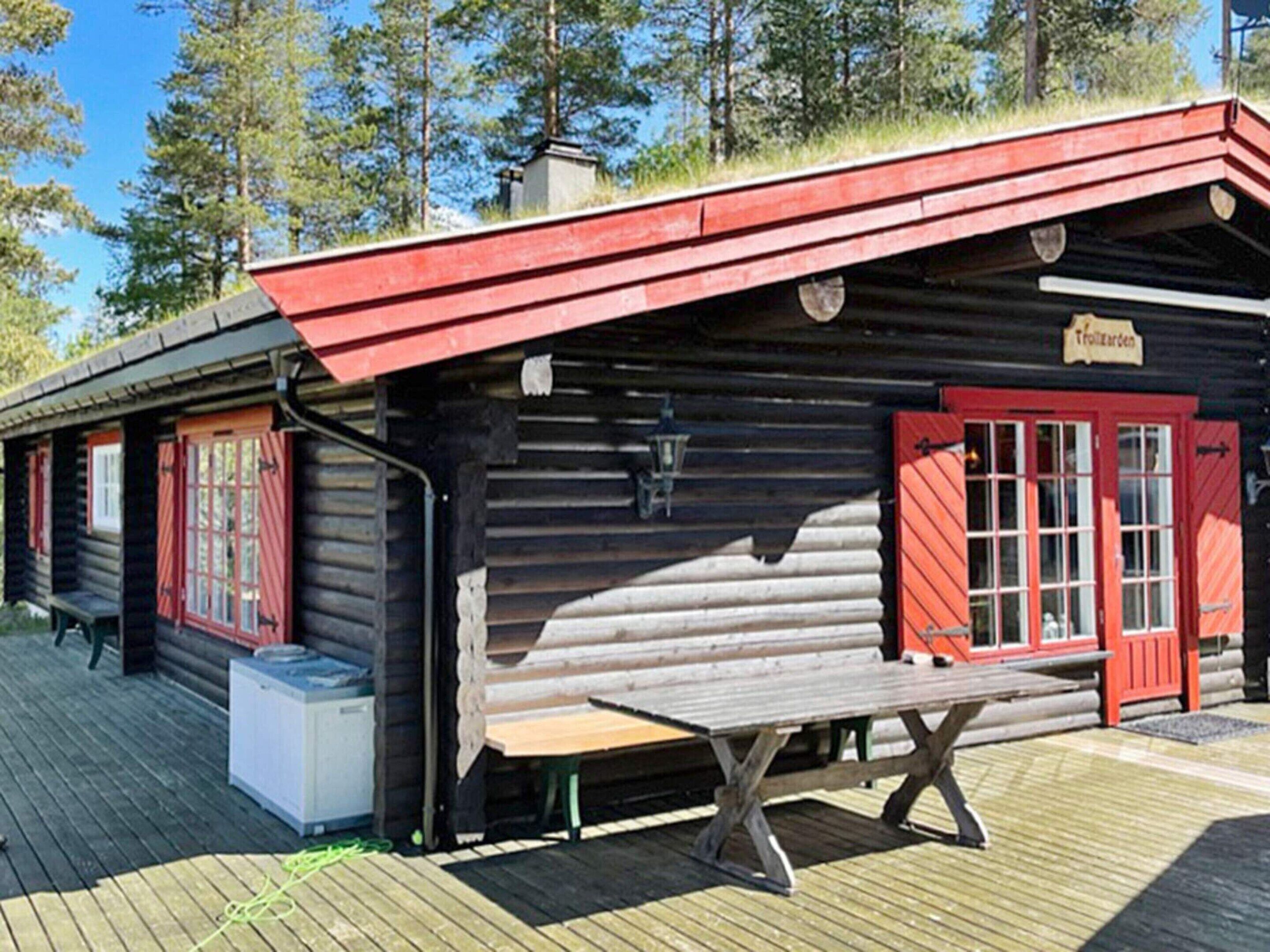 A log cabin with a red door and windows, a bench outside, and a sign that reads "Trollgard".