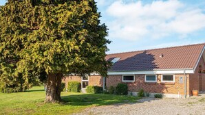 A single-story brick house with a sloped roof, a gravel driveway, and a large tree in the foreground.