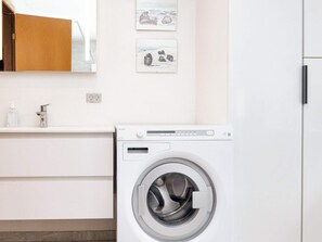 A modern laundry room with a front-loading washing machine, a white sink with a faucet, and a wooden cabinet above the sink.