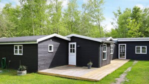 A row of dark-colored cabins with a wooden deck in front, surrounded by green grass and trees. - 4 Star Holiday Home in Vig (Vig)