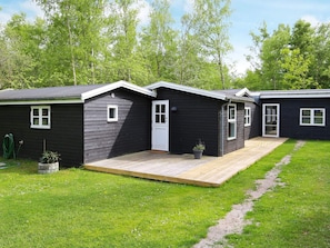 A row of dark-colored cabins with a wooden deck in front, surrounded by green grass and trees. - 4 Star Holiday Home in Vig (Vig)