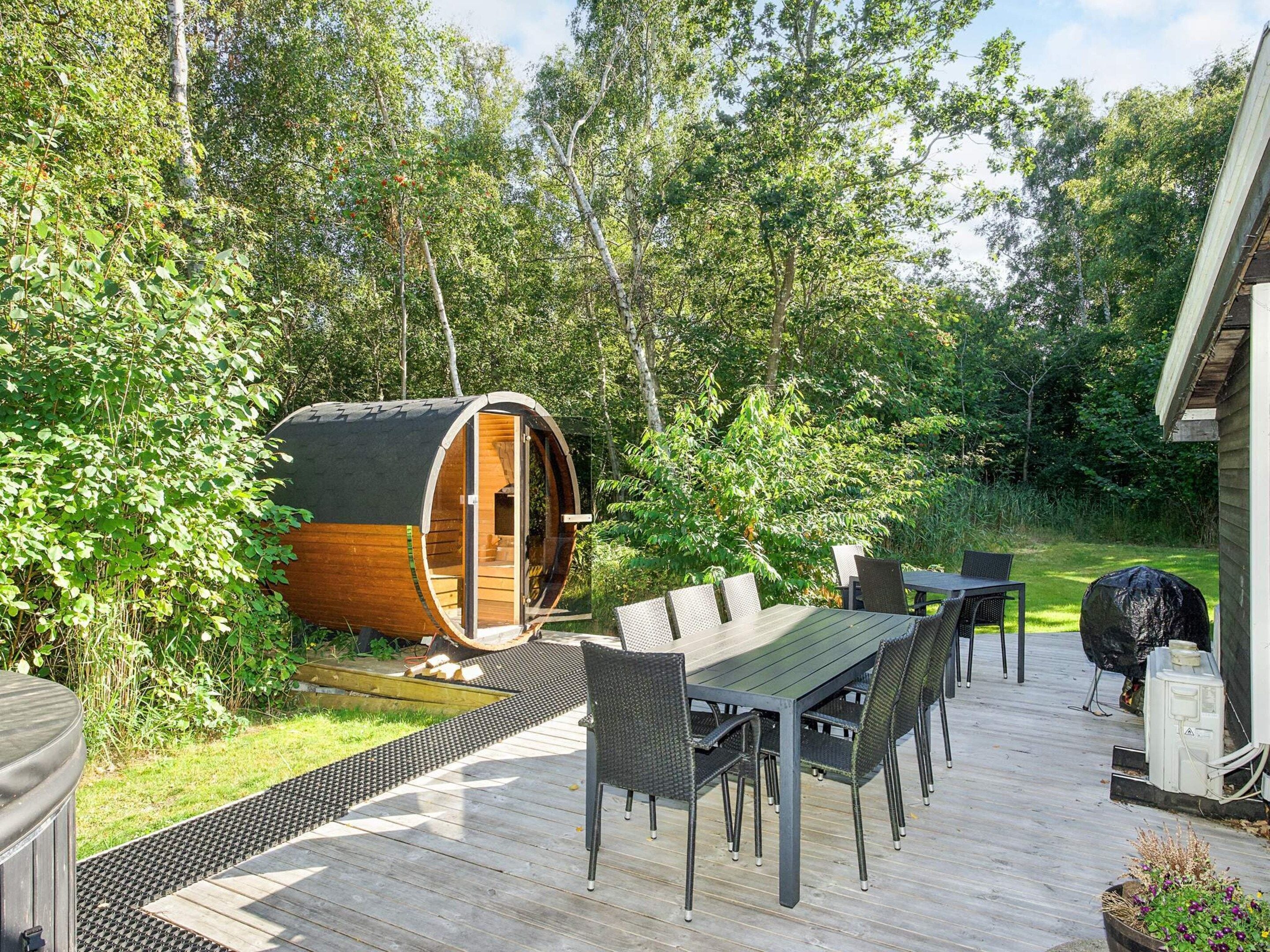 A wooden deck with a dining table and chairs, a sauna, and a large barrel-shaped structure in a forested area.