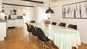 A dining area with a long table, chairs, a hanging pendant light, and a painting on the wall.