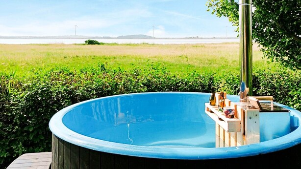A hot tub with a wooden exterior and a metal pole, surrounded by greenery and a view of a body of water. - 4 Person Holiday Home in Fjerritslev (Fjerritslev)
