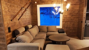 Une pièce chaleureuse au décor intérieur en bois, une grande fenêtre donnant sur un paysage enneigé, un canapé, une petite table et un porte-skis fixé au mur.