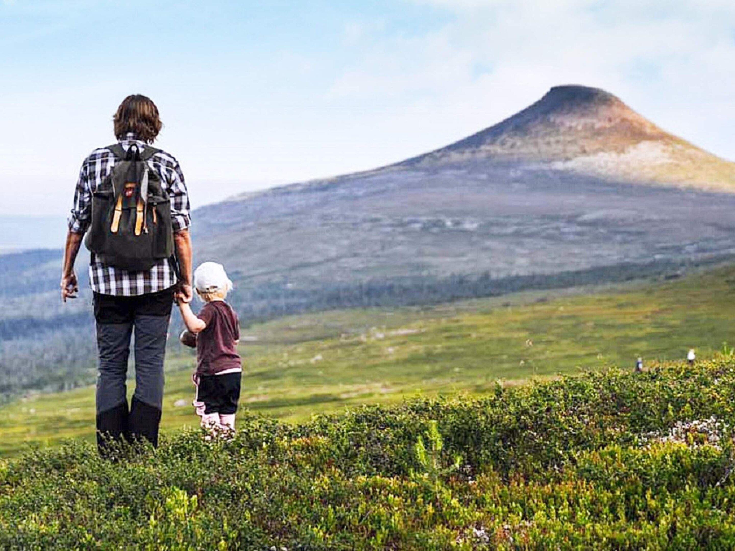 A person walking with a child on a grassy hill with a mountain in the background.