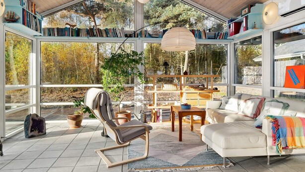 A spacious room with a high ceiling, large windows, and a wooden ceiling. There is a white sofa, a chair, and a small table with a bowl. Shelves with books and decorative items are visible. - 8 Person Holiday Home in Hadsund (Hadsund)