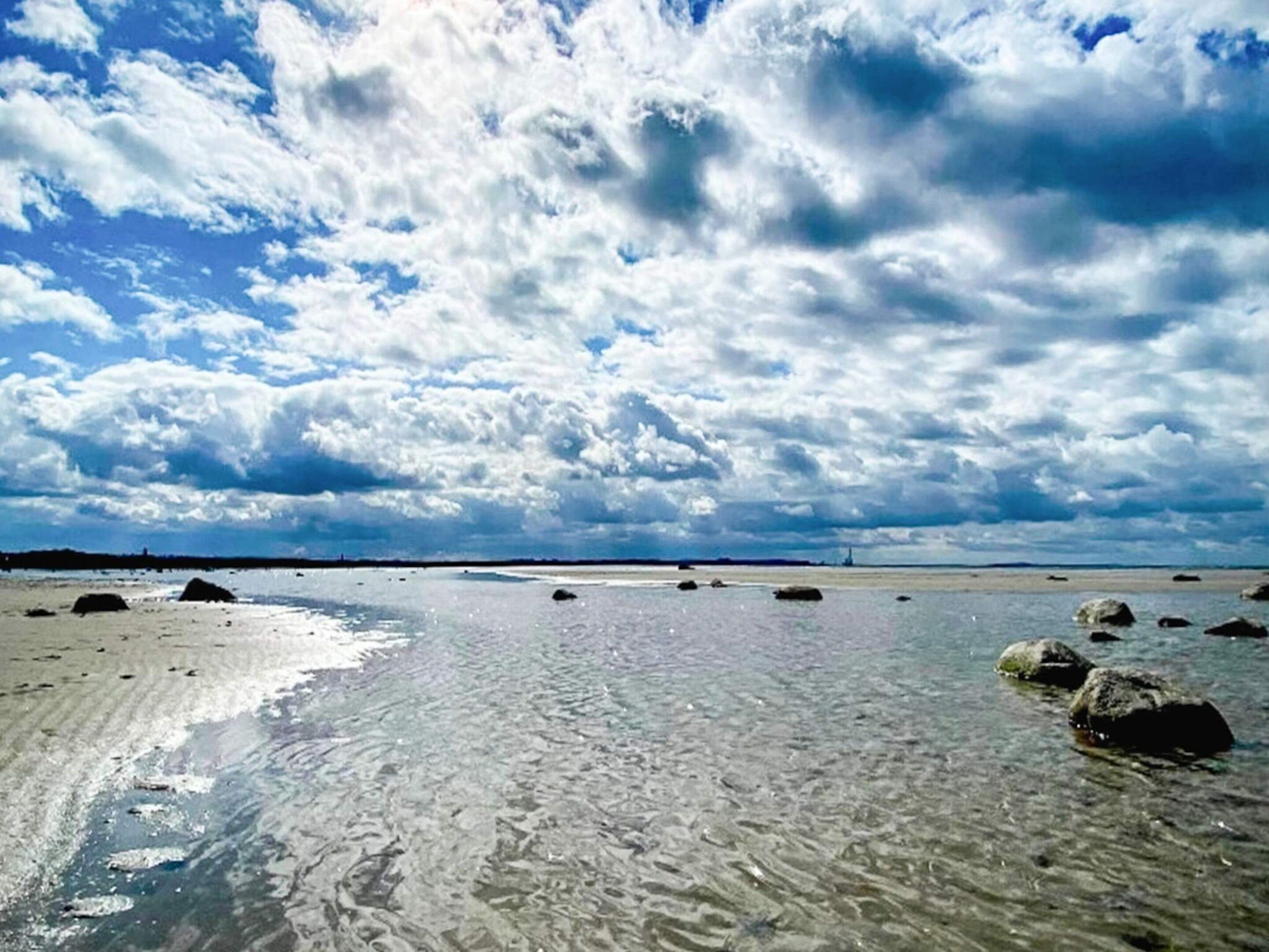En strand med klart vatten, klippor och en molnig himmel.