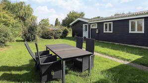 Ein kleiner, dunkel gefärbter Wohnwagen mit einer Tür und zwei Fenstern, einem grünen Rasen und einem Tisch mit Stühlen.