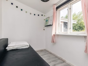 A small, neatly arranged bedroom with a single bed, a window with curtains, and a wall-mounted shelf with a framed picture.