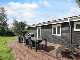 Una terraza de madera con mesa y sillas, una parrilla y una casa de exterior oscuro con detalles blancos.