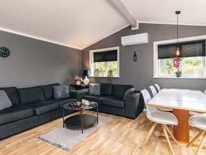 A modern living room with a dark grey sofa, a round coffee table, and a dining table with chairs. There are two windows with blinds, a wall-mounted air conditioner, and a hanging light fixture. - 4 Star Holiday Home in Jaegerspris (Jægerspris)