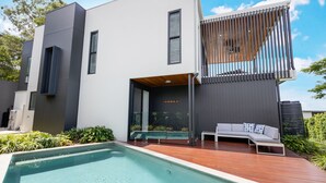Ett modernt hus med en swimmingpool, ett trädäck och en inbyggd utomhusplats med sittmöbler.