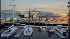 En småbåtshamn med flera båtar förtöjda, en restaurang vid vattnet och en modern byggnad med ett sluttande tak.