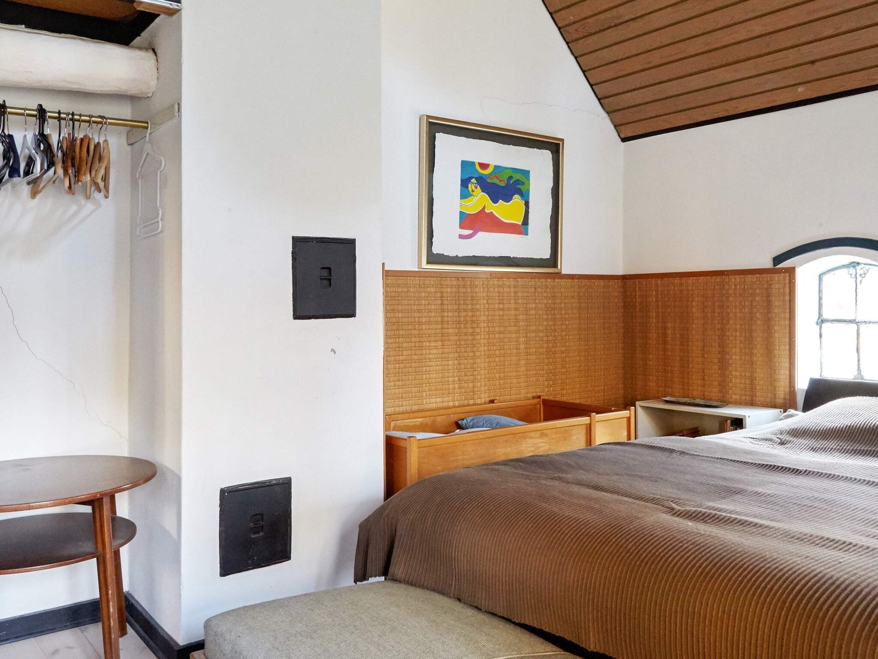 A bedroom with a bed, a bedside table, a chair, a window, and a wall-mounted light switch.